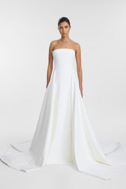 A model wearing the Mati Gown, a floor-length ivory bonded crepe strapless wedding dress with a straight neckline, contrast satin band, side split, and a dramatic train.