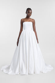 A model wearing the Cleo Gown, a floor-length ivory strapless wedding dress featuring a straight neckline, a low-dipped back, and a tightly gathered full skirt with a sweeping train.