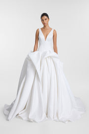 A model wearing the Bailey Gown, a floor-length ivory duchess satin ballgown featuring a fitted bodice with a deep V-neckline, waist draping, and a voluminous skirt with sculptural folds and a sweeping train.